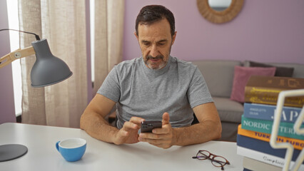 Canvas Print - Middle-aged man sitting indoors in a living room, using smartphone, with books and a lamp on a table