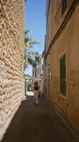 Fototapeta Uliczki - Young woman walking in the narrow, sunlit streets of fornalutx, mallorca, spain, surrounded by traditional mediterranean architecture and vibrant greenery.