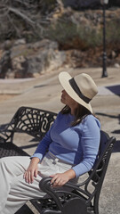 Wall Mural - Young hispanic woman sits relaxed in an urban park, exuding elegance with her fashionable hat and casual pose