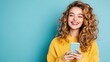 © Sumet - Happy woman with curly hair holds a smartphone, wearing a bright yellow sweater against a blue background, radiating joy and positivity.