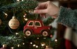 © Georgii - A hand decorates a Christmas tree with a red car ornament amid festive lights and traditional baubles