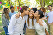 © luisrojasstock - A woman smiles brightly as two friends kiss her on the cheeks during an outdoor gathering, surrounded by other happy friends.