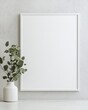 © Naphol - Minimalist decor with a blank picture frame and a vase of green leaves.