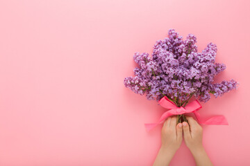 Little baby girl hands holding fresh beautiful flower bouquet of purple lilac with ribbon on pink table background. Pastel color. Closeup. Point of view shot. Empty place for text. Top down view.