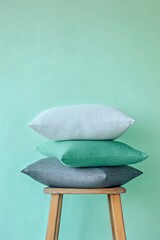 Three decorative pillows of different colors are stacked on a wooden stool against a mint green wall.