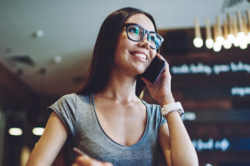 Cheerful female teenager communicate with birthday organisation service via cellular phone for planning party, happy hipster girl calling to friend for making positive telephone conversation