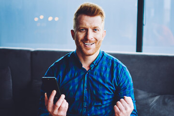 Portrait of happy emotional male blogger feeling excited from statistics about own web page in social networks, prosperous hipster guy with modern smartphone gadget in hand looking at camera