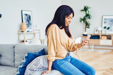 Young African American millennial woman sitting at sofa in living room using mobile phone connected to 4G at free time, dark skinned hipster girl share content in social networks on cellular