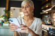 © Marko Geber - Senior woman using smartphone at home kitchen