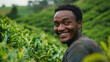 © Anastasiia Havelia - A happy young African man working in tea plantations, picking fresh leaves and smiling at the camera, with lush green fields of tea bushes behind him. The focus is on his face show