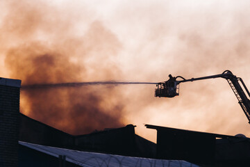 Wall Mural - Firefighters use telescopic tower during massive large blaze fire, firetruck with crane, firemen team putting out the fire with extendable fire ladder, blazing warehouse factory, burning building