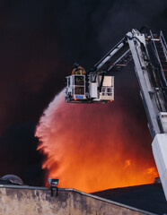 Wall Mural - Firefighters use telescopic tower during massive large blaze fire, firetruck with crane, firemen team putting out the fire with extendable fire ladder, blazing warehouse factory, burning building