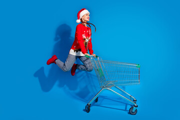 Wall Mural - Full body photo of attractive young woman push shopping cart jumping dressed red christmas clothes theme party on blue color background