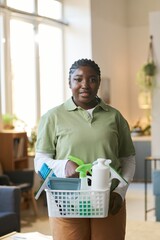 Wall Mural - Vertical waist up portrait of smiling African American woman working in cleaning service and looking at camera indoors while holding box with supplies