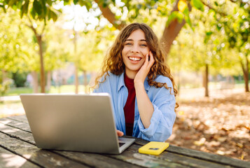 Wall Mural - Young woman brought her laptop to the park to get some fresh air and work. Online education, Freelance work, technology concept.