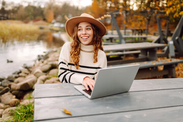 Wall Mural - Working surrounded by autumn nature. Young woman with laptop at a rustic table near a tranquil waterway in autumn. E-learning concept. Remote work.