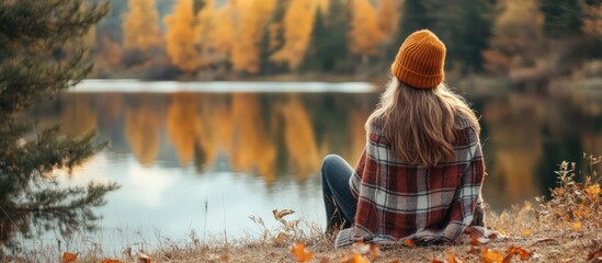 Canvas Print - Woman in a knitted hat and plaid blanket sits on a lakeshore and admires the fall foliage reflection in the water.