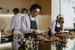 © AnnaStills - Professional cooking team in white uniform with black aprons preparing ingredients for caprese appetizer, African American female cook speaking on phone with client and writing down details
