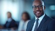 © BoOm - Smiling businessman in a suit with glasses, blurred colleagues in the background, professional office setting.