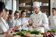 © tynza - A chef teaching a cooking class, surrounded by students, explaining a technique with ingredients on the table, educational and professional environment.