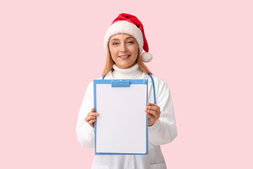 Poster - Mature female doctor in Santa hat with clipboard on pink background