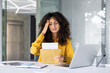 © Liubomir - Stressed woman receiving bad news from envelope in office setting. Expression of worry while seated at desk with laptop, phone, and various office essentials. Modern professional environment.