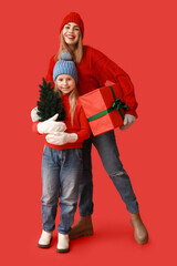 Poster - Cute little girl with small Christmas tree and her mother holding gift box on red background