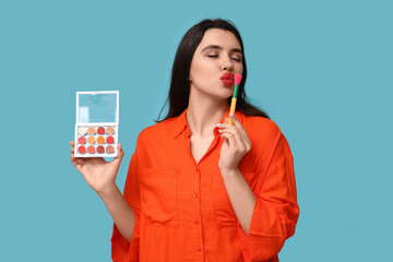 Sticker - Beautiful young woman with palette of eyeshadows kissing makeup brush on blue background