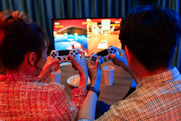 Couple gamer with controlled joystick playing car racing video game together on tv screen with snack at back side view, sitting sofa at neon light colorful living room at modern comfy home. Infobahn.