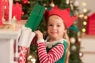 Sticker - Cute little girl in elf costume taking gift from sock at home on Christmas eve, closeup