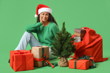 Poster - Beautiful young happy African-American woman with Christmas tree, Santa bag and gift boxes sitting on green background