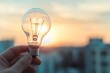 © Sebelas Studio - Hand holding a light bulb against a blurred cityscape at sunset, symbolizing innovative ideas and energy