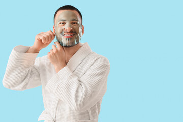 Canvas Print - Handsome young happy man in bathrobe applying cosmetic gel mask on face against blue background