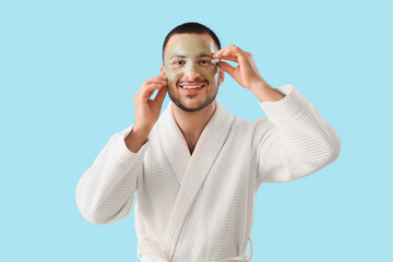 Canvas Print - Handsome young happy man in bathrobe applying cosmetic gel mask on face against blue background