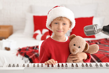 Sticker - Cute little boy in Santa hat with toy bear and synthesizer at home on Christmas eve