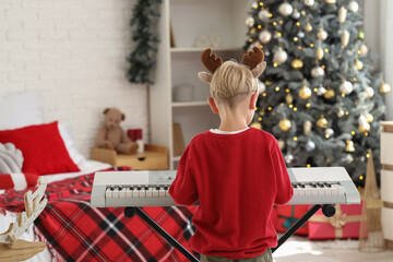 Sticker - Cute little boy in reindeer horns playing synthesizer at home on Christmas eve, back view