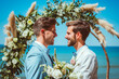 © MVProductions - Handsome gay couple in wedding ceremony at outdoor venue near sea under wedding flower arch, Authentic LGBTQ Relationship.