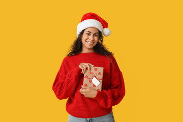 Wall Mural - Happy African-American woman in Santa hat and with Christmas gift on yellow background