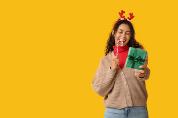 Wall Mural - Happy African-American woman with Christmas gift and candy cane on yellow background