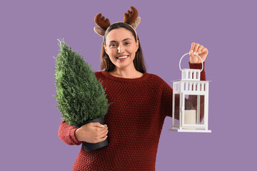 Wall Mural - Young woman in reindeer horns with Christmas tree in pot and lantern on purple background