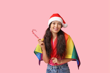 Poster - Young Asian woman in Santa hat with LGBT flag and candy cane on pink background. Christmas celebration