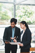© Satori Studio - Two business professionals in formal attire discussing documents in a modern office with large windows and natural light.