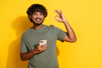 Poster - Photo of good mood guy with wavy hair dressed khaki t-shirt hold smartphone showing okey approve isolated on yellow color background