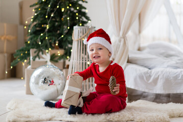 Poster - Smiling little baby boy in a santa costume and red hat sitting on a soft fur carpet in front of a christmas tree