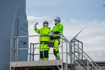 Maintenance engineer team standing at windmills at wind turbine farm. Group of people wear safety helmet and uniform working at alternative renewable energy wind station. Sustainable energy technology