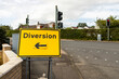 © Tosh - Yellow diversion sign with arrow pointing left on metal frame at road junction intersection with traffic lights, brick wall, and hedges in background