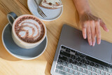 Close-up of hand on laptop with coffee break essentials on wooden table. Close-up of hand on laptop with cocoa and dessert on wooden table