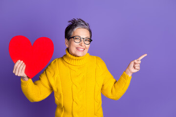 Poster - Photo of charming positive woman wear yellow sweater holding heart pointing empty space isolated violet color background