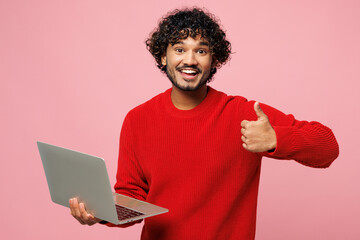 Poster - Young cheerful smart IT Indian man wear red sweater casual clothes hold use work on laptop pc computer show thumb up isolated on plain pastel light pink background studio portrait. Lifestyle concept.