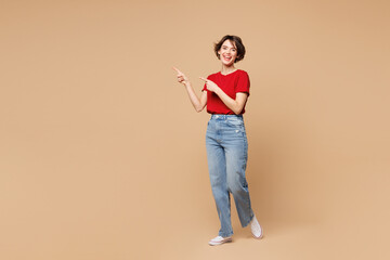 Full body young smiling fun happy woman she wear red t-shirt casual clothes point index finger aside on area mockup isolated on plain pastel light beige background studio portrait. Lifestyle concept.